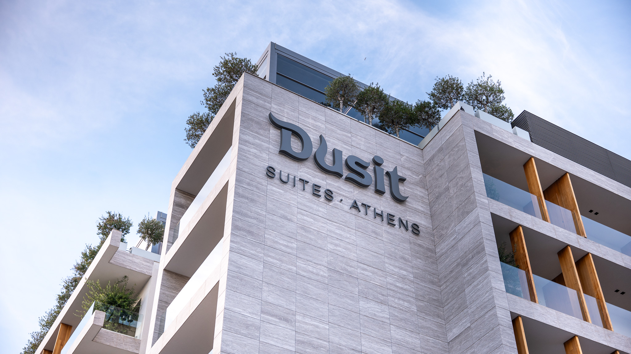 Dusit Suites Athens, low-angle facade with Dusit signage and rooftop tree line